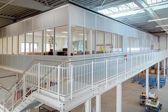 Case-Study-Kleusberg-Architecture-Modular-Construction Modern industrial hall with elevated modular office area, white steel structure and staircase; underneath are work and technical areas.