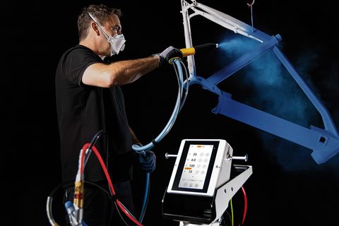Operator performing electrostatic powder coating on a bicycle frame using a Gema powder coating gun OptiSelect ProX and OptiStar Pro X control unit to achieve uniform coating quality and efficient process control.